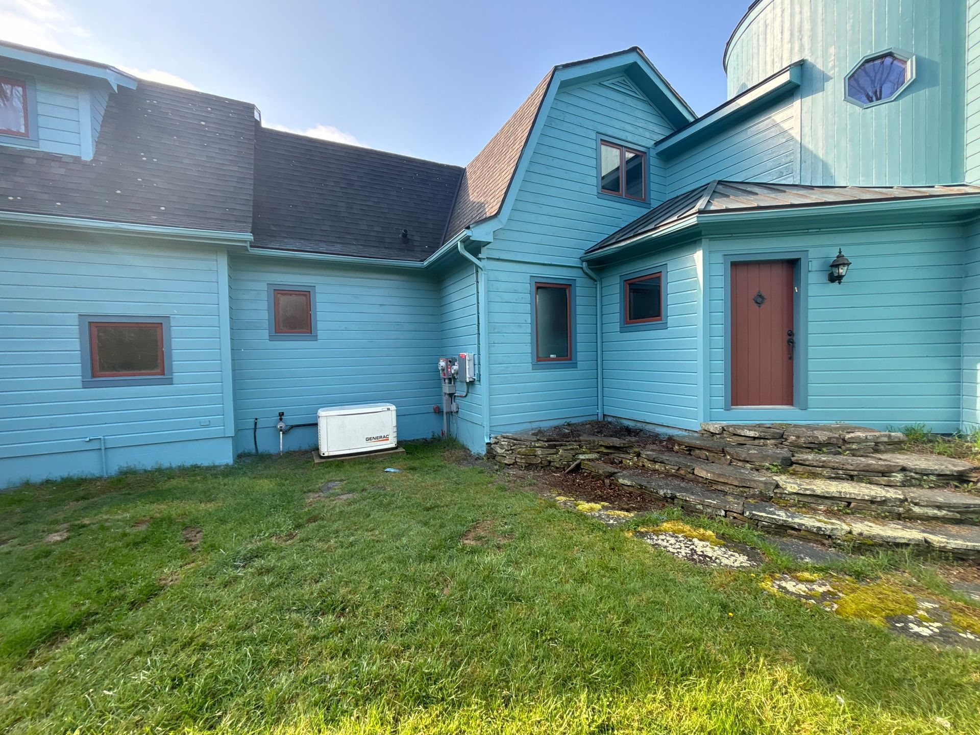 A light blue house with a dark roof and a red front door, featuring a stone patio and a grassy lawn in the foreground.