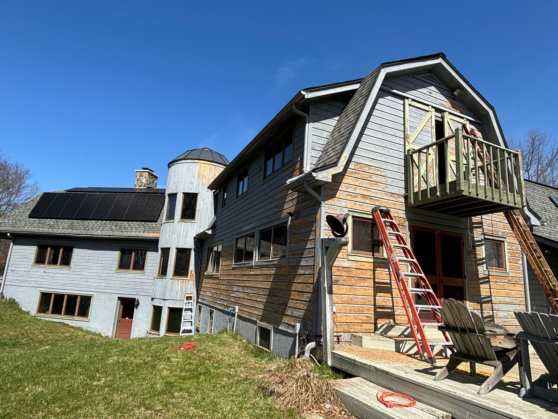 A two-story grey farmhouse with a silo, solar panels, and a wooden balcony under construction, with a ladder leaning on it.
