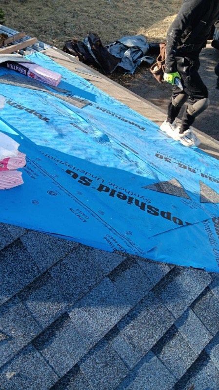 A worker in protective clothing installs blue roofing underlayment on a shingled roof.