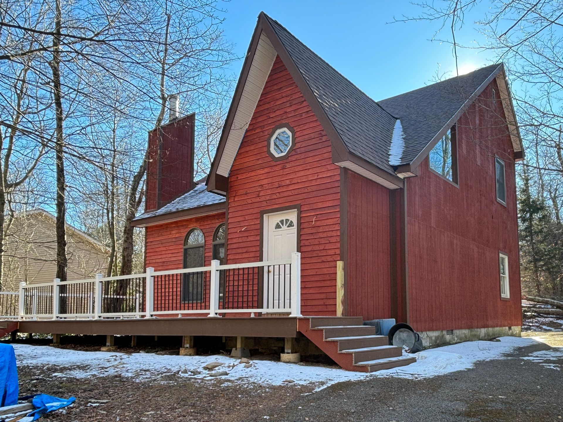 A two-story red cabin in a wooded area with a white porch, brown roof, and patches of snow on the ground.