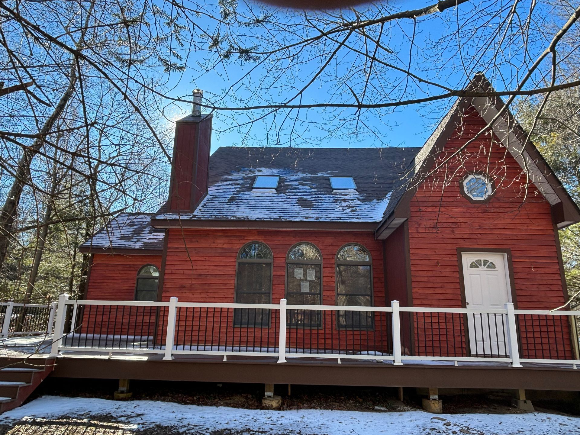 A red, two-story cabin with a deck, a white door, and arched windows, surrounded by trees dusted with snow.