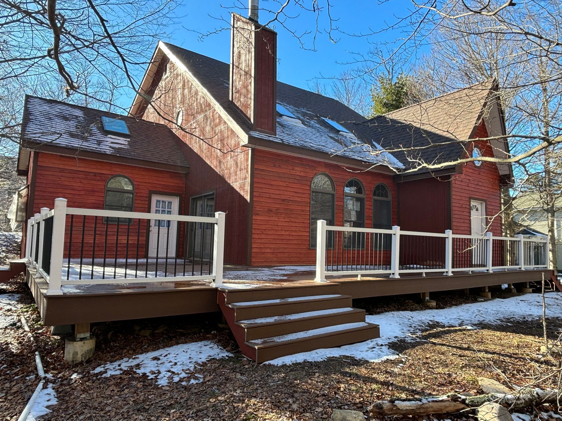 A two-story red home with a large deck, white railing, and wooden stairs in a snowy, wooded setting under a clear blue sky.