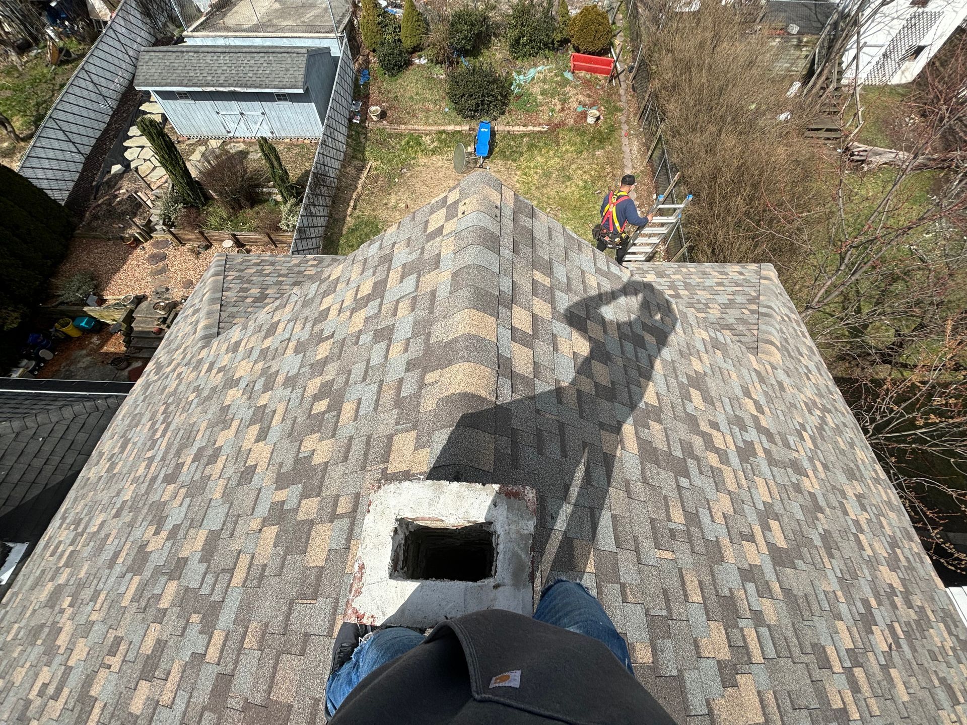 A high-angle perspective from a roof, showing a chimney, shingles, and a person standing on a ladder in the yard below.