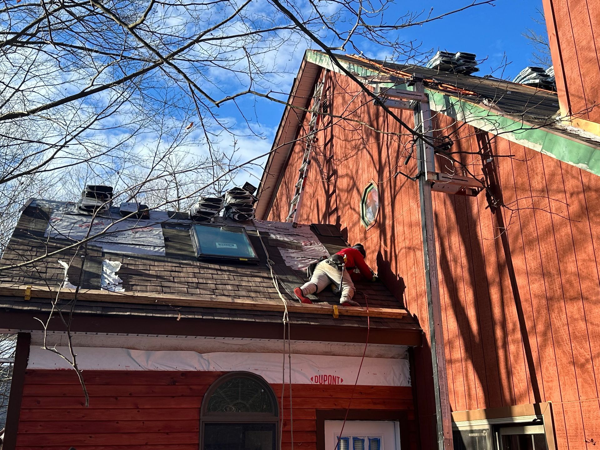 A person in a red shirt works on a partially stripped roof of a red house under a bright blue sky.