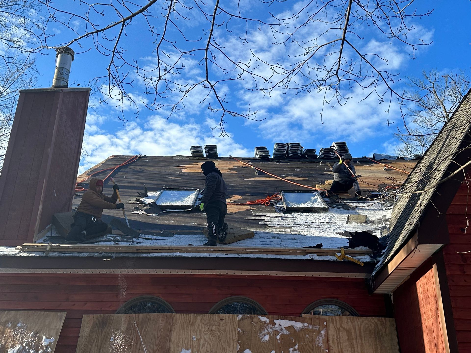 Three workers perform roof repairs on a red-sided house against a sunny, blue sky.