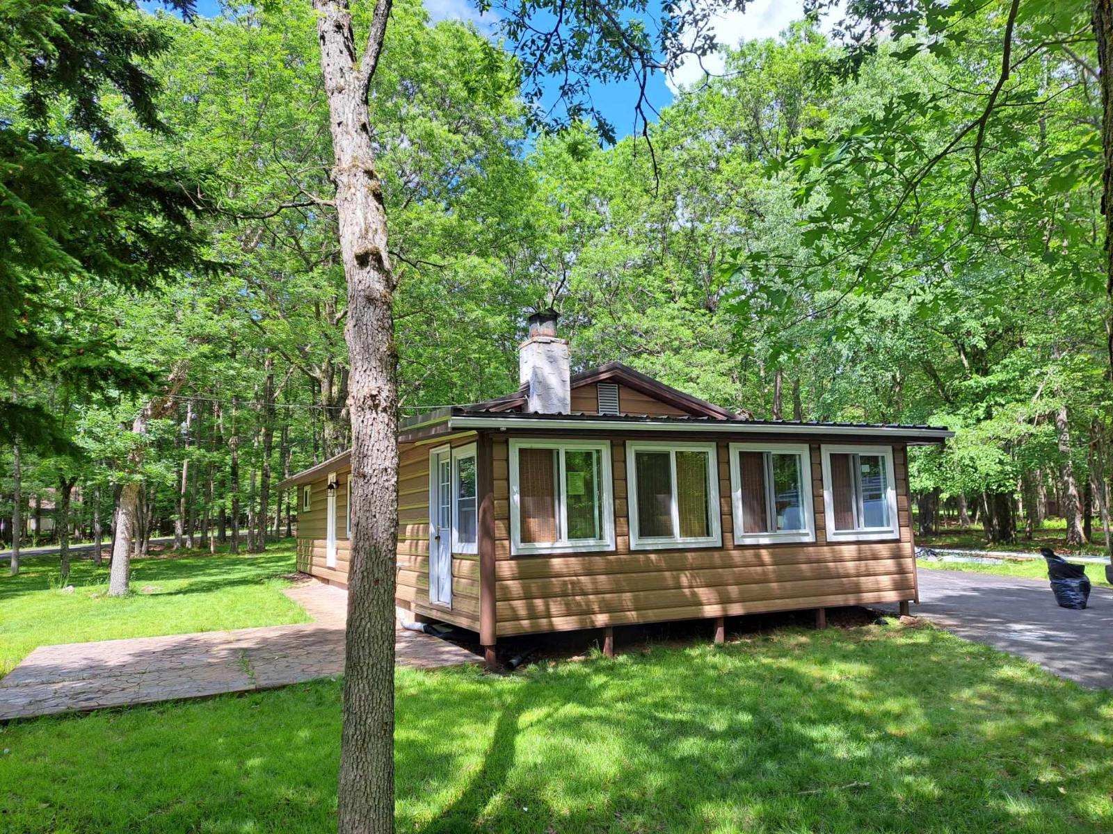 A single-story log cabin with a white chimney sits in a lush, green forest on a sunny day.