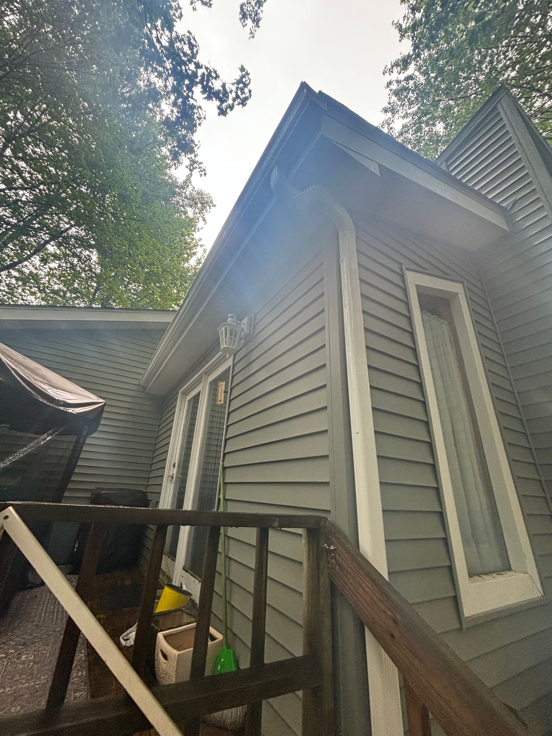 Exterior of a gray-sided house showing a wooden deck railing, a window, and a white downspout under a slanted roof.
