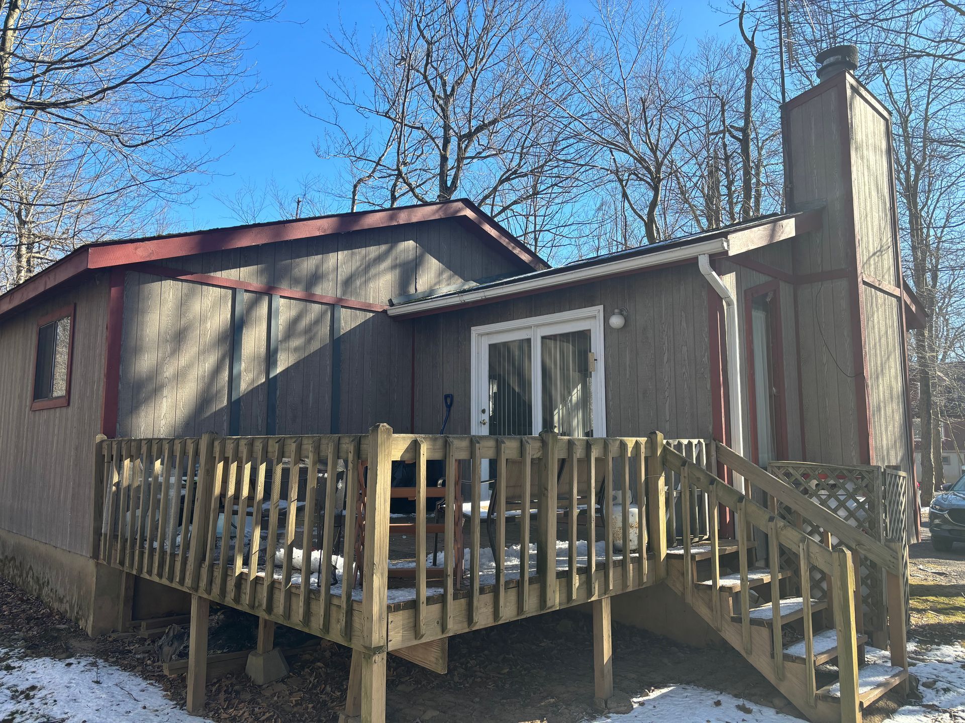 A small, brown, single-story cabin with a wooden deck and exterior chimney, surrounded by trees on a snowy day.