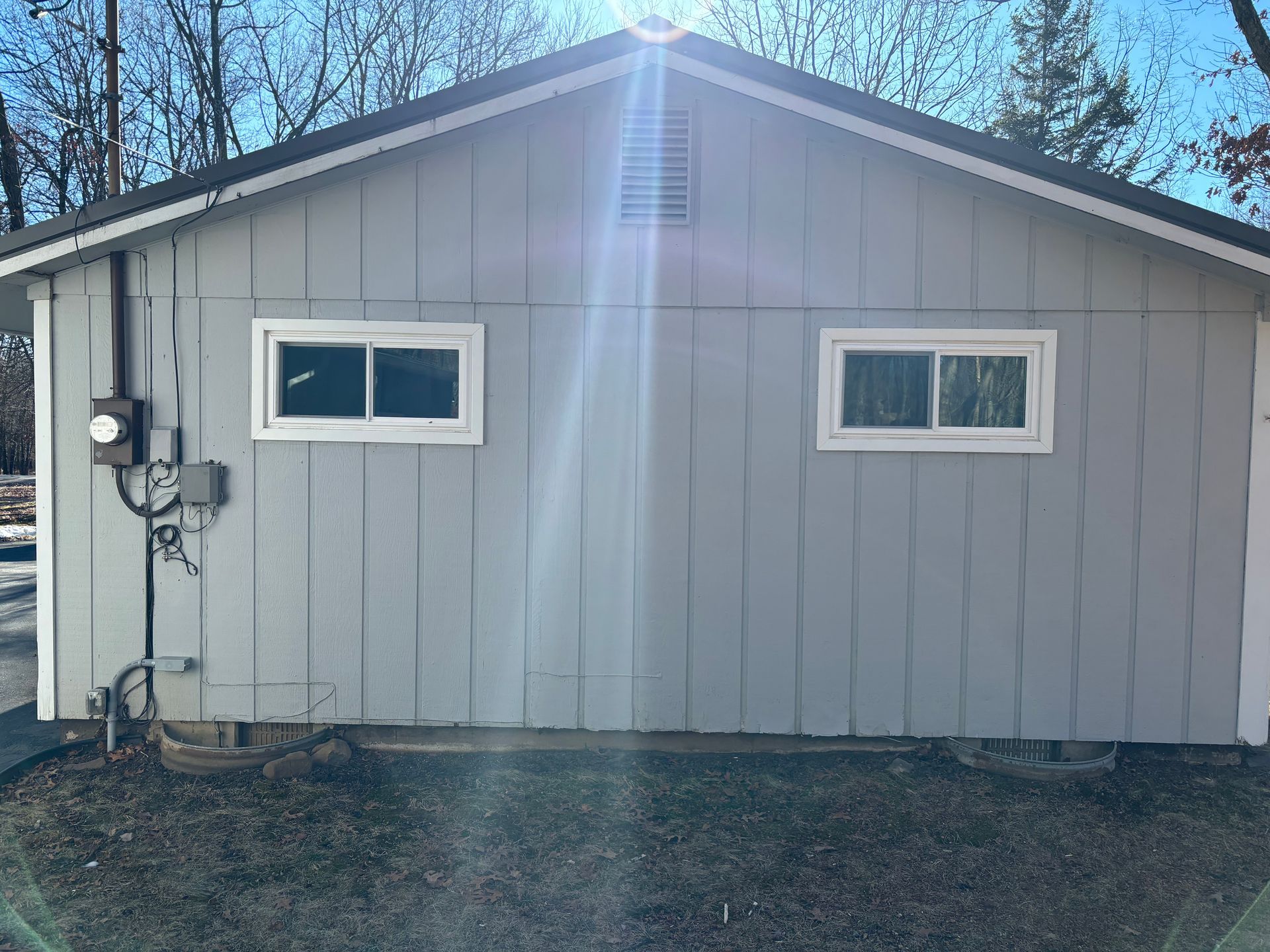 A gray, rectangular garage with vertical siding, two white-framed windows, and an electrical meter on the left wall.