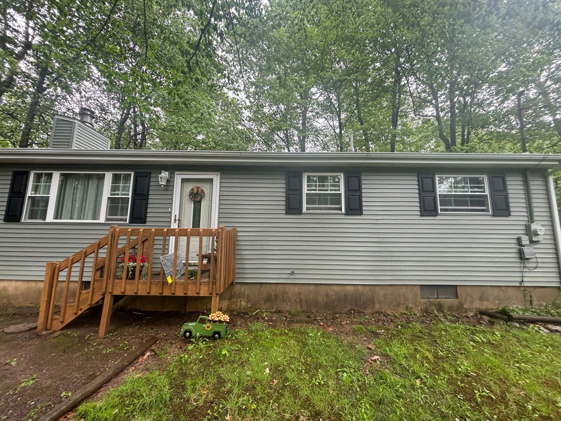 A gray, single-story house with horizontal siding, dark shutters, and a wooden front porch, situated in a wooded area.