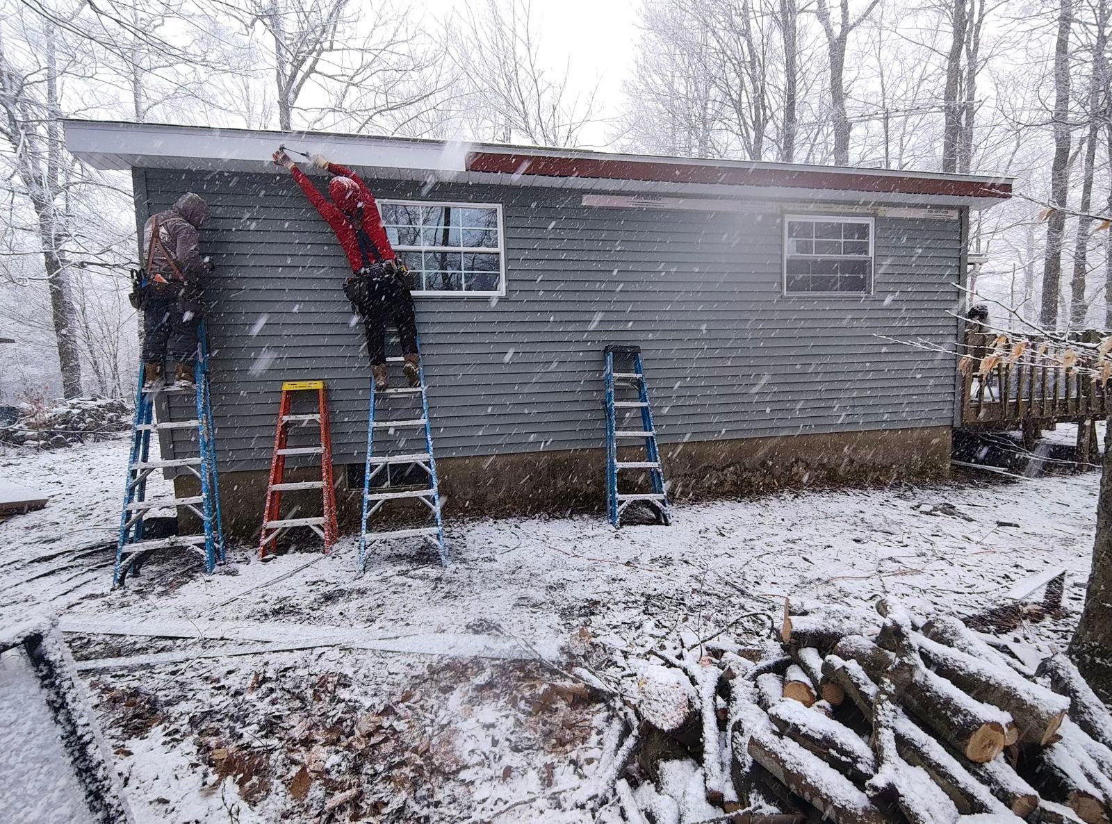 Two people on ladders work on the roofline of a grey siding house during a snowstorm, with logs in the foreground.
