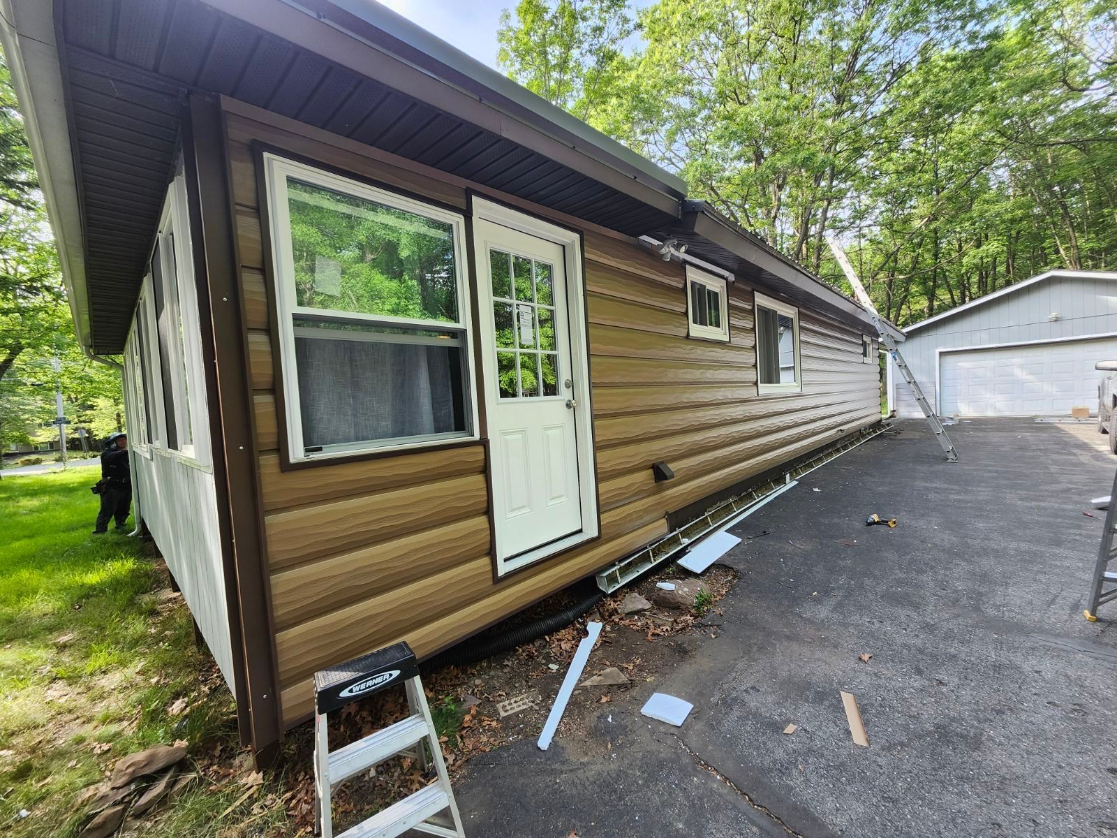 A tan log-style cabin with a white door and windows, situated next to a paved driveway and a detached garage.