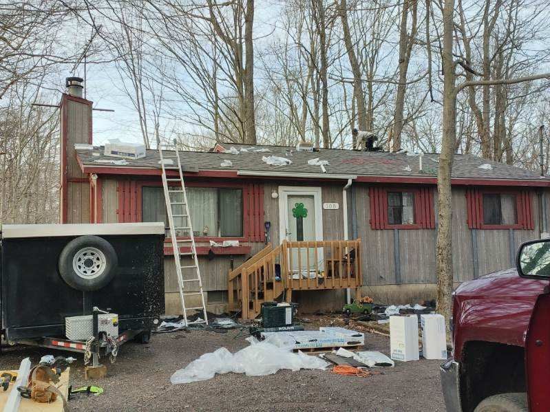 A one-story house under renovation with a ladder against the roof, a parked trailer, and building materials in the yard.
