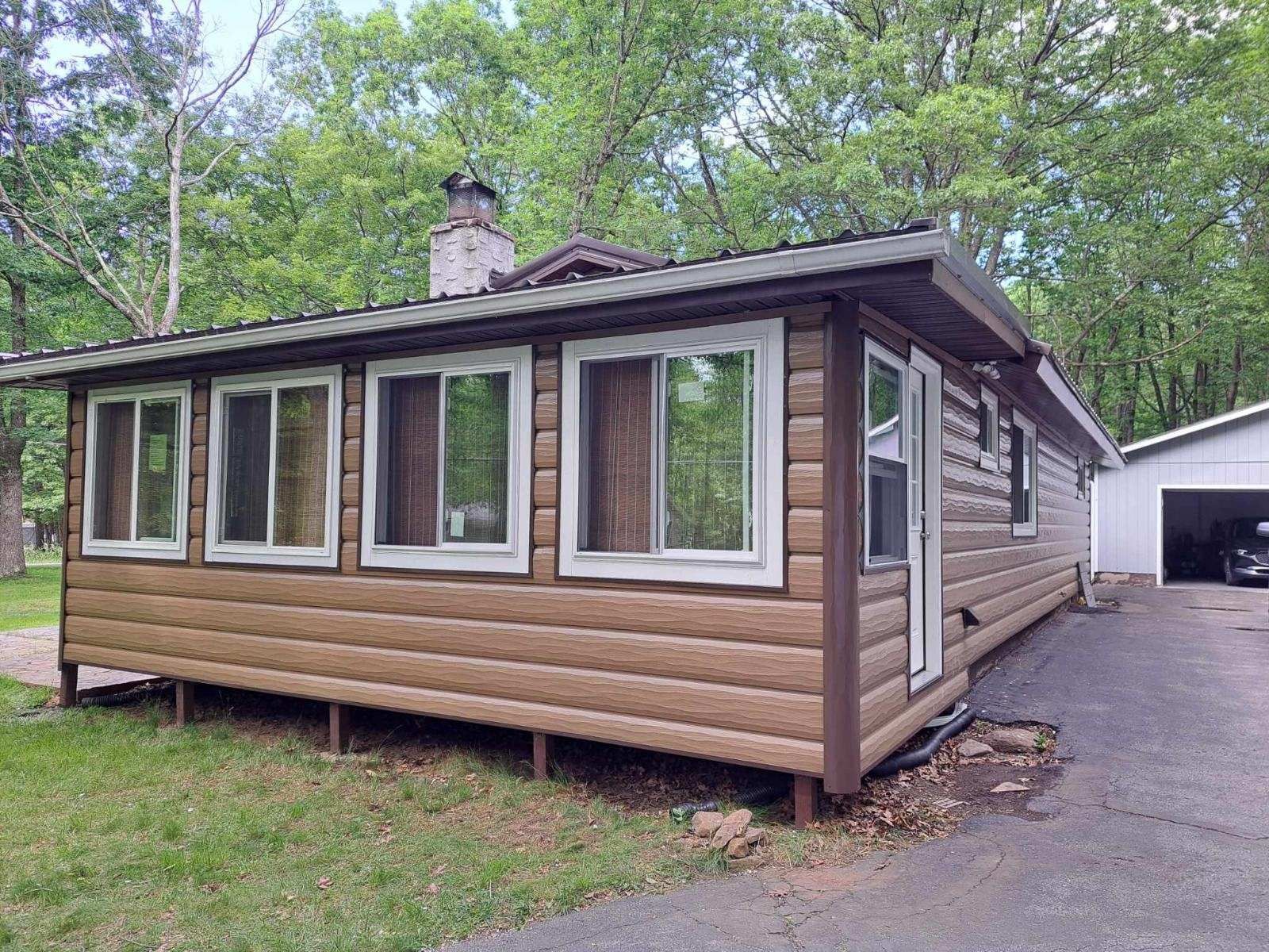 A brown log-sided cabin with white-framed windows sits on a grassy lot next to a gravel driveway and a white garage.