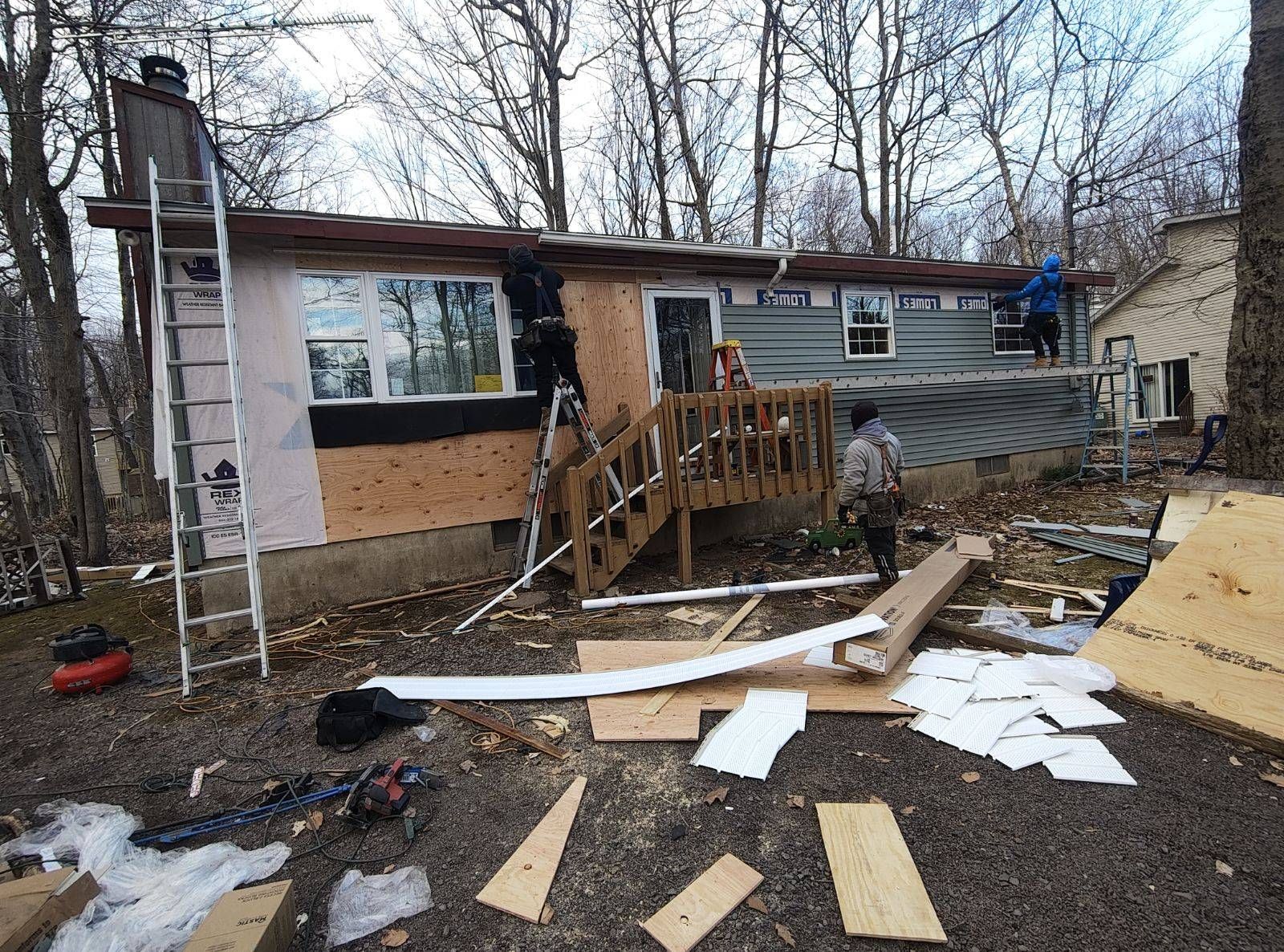 Three workers on ladders and scaffolding renovate the exterior of a house, installing new siding and wood paneling.