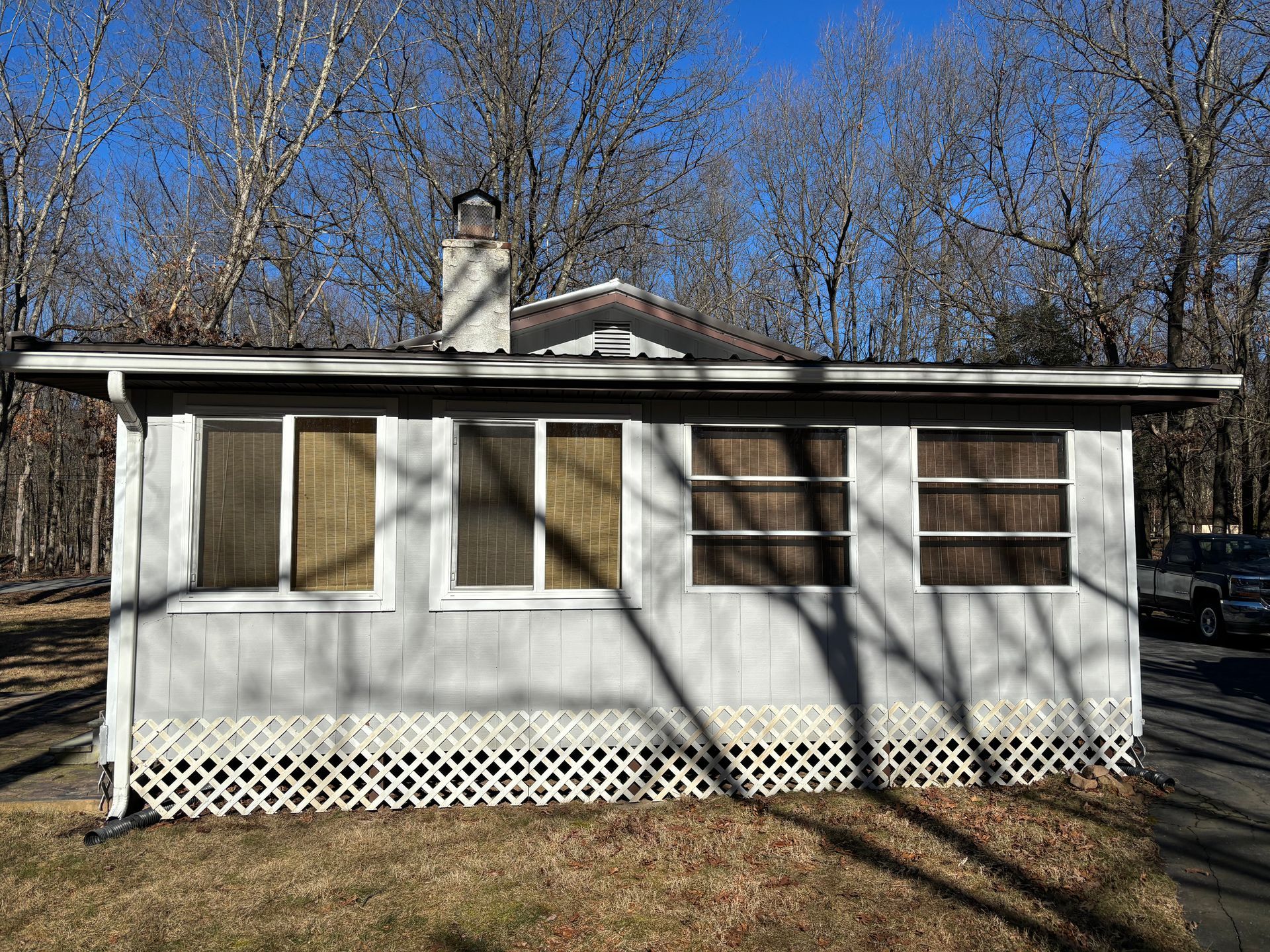 Single-story light gray house with white lattice skirting, a brick chimney, and windows covered by tan blinds in a woods.