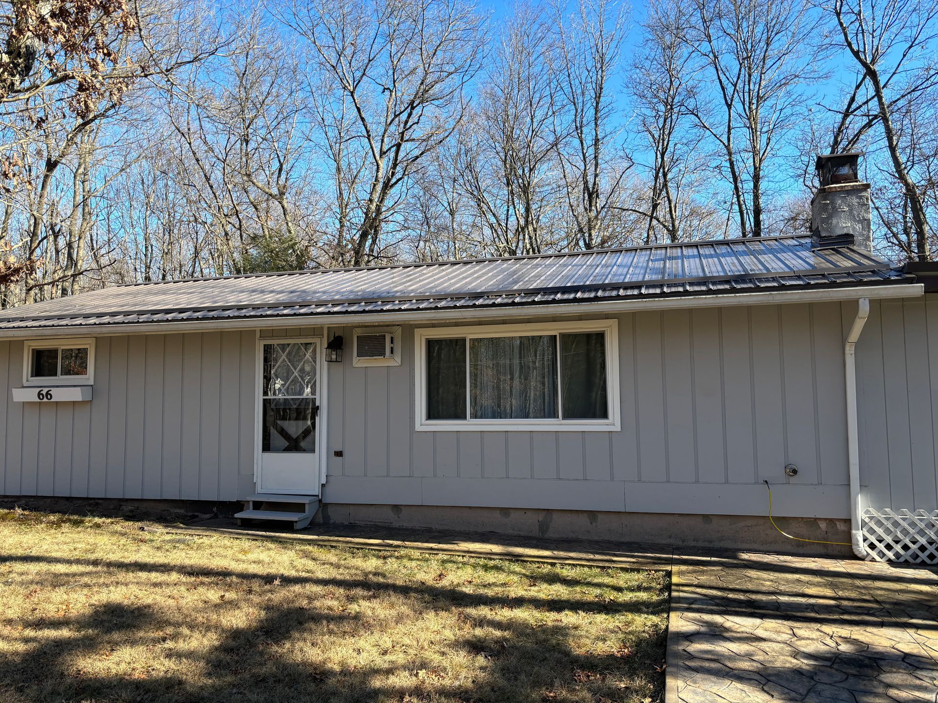 A light gray, one-story house with a metal roof and a brick chimney stands among bare trees on a sunny day.