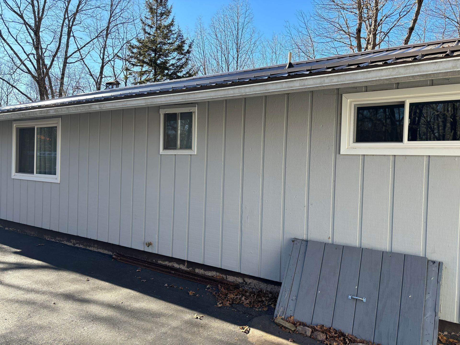 A side view of a light gray, vertical wood-sided house with three white-framed windows against a blue sky.