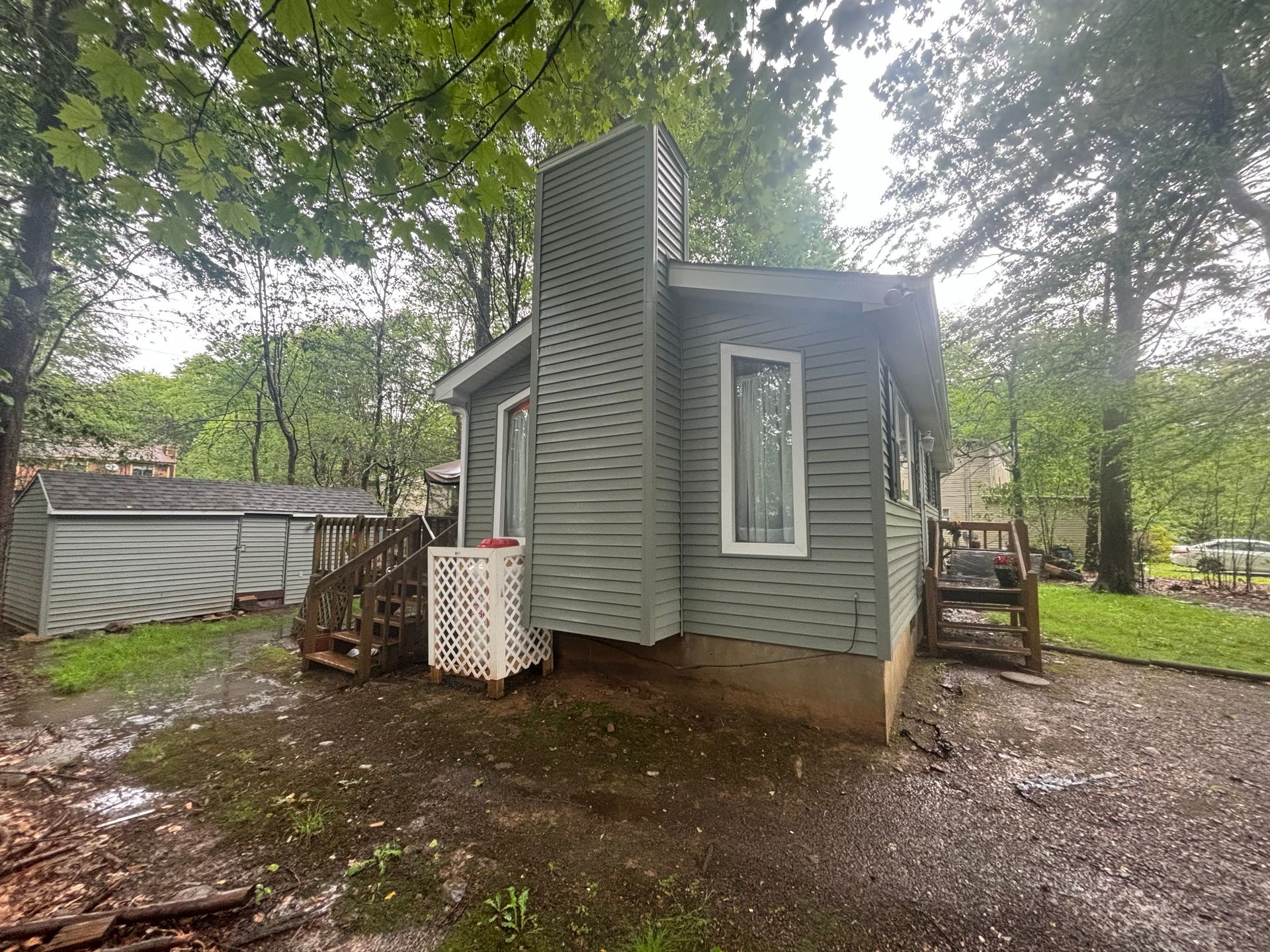 A gray one-story house with a prominent chimney and white lattice skirt, set in a wooded area with a small side porch.