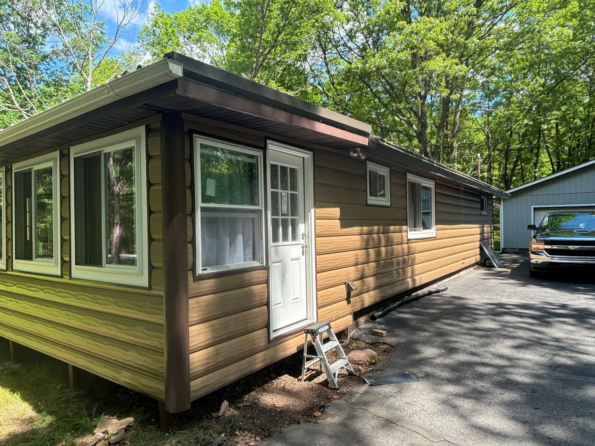 A tan log-style cabin with white trim and a white door, situated in a wooded area next to a gravel driveway.