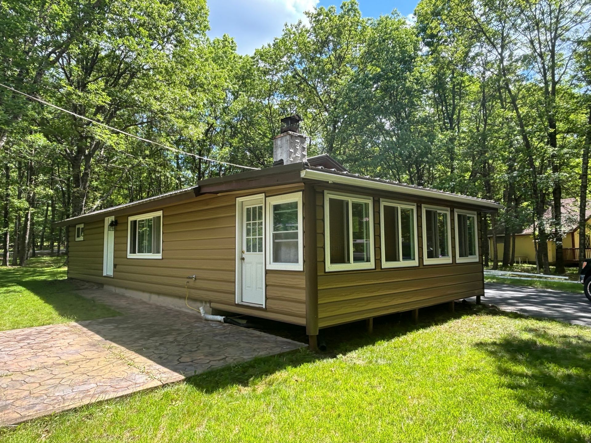 A tan, single-story cabin with a metal roof and a small stone chimney, nestled in a lush, wooded area.