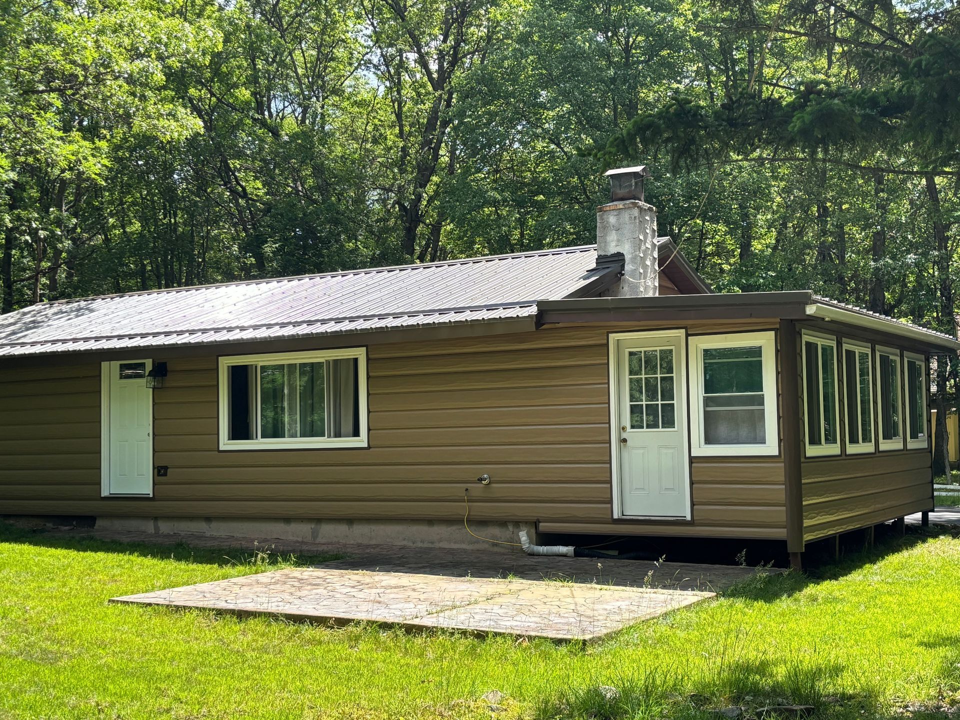 A brown cabin with a metal roof and stone chimney sits in a grassy clearing surrounded by dense trees.