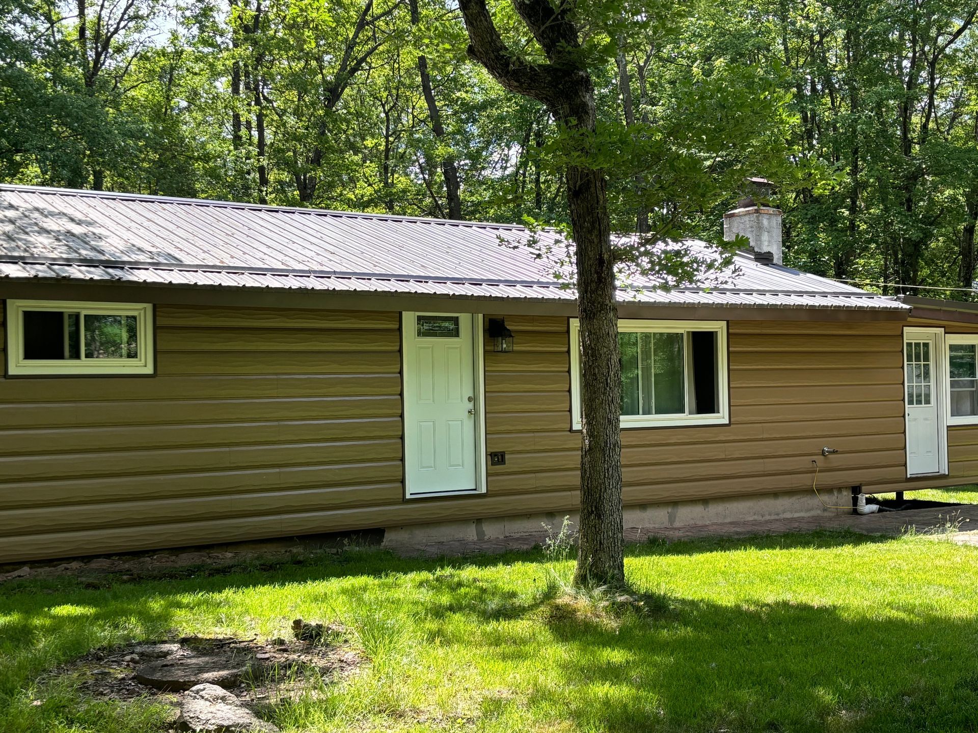 A single-story brown cabin with a metal roof and white doors, surrounded by trees and a green lawn.