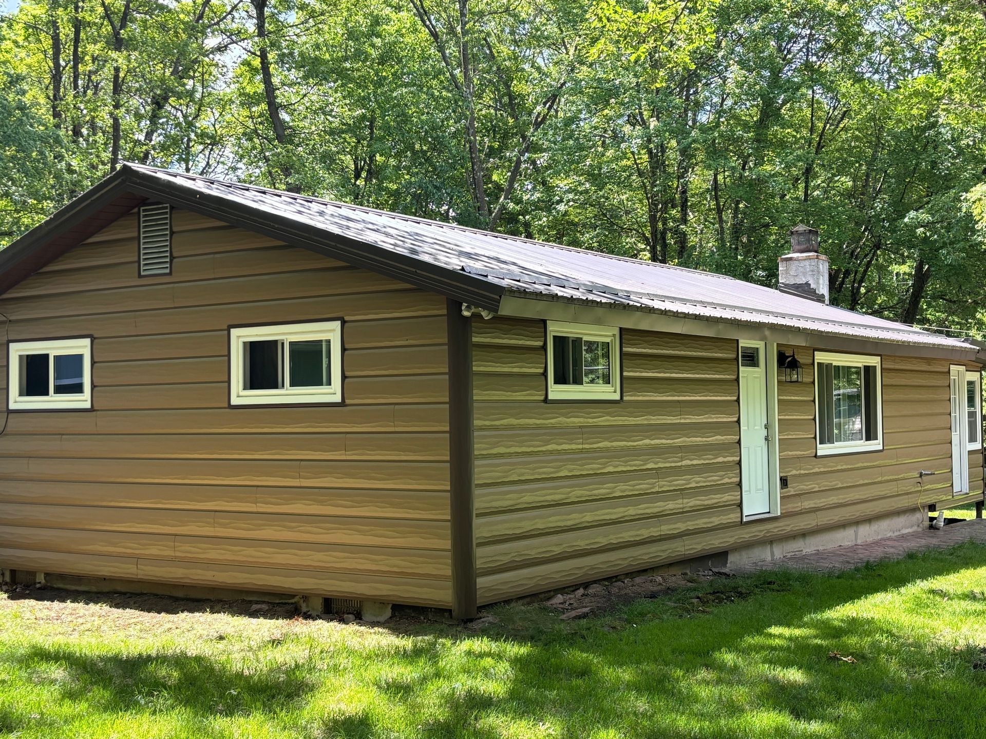 A brown cabin with white trim, a metal roof, and a chimney, set in a yard surrounded by green trees.