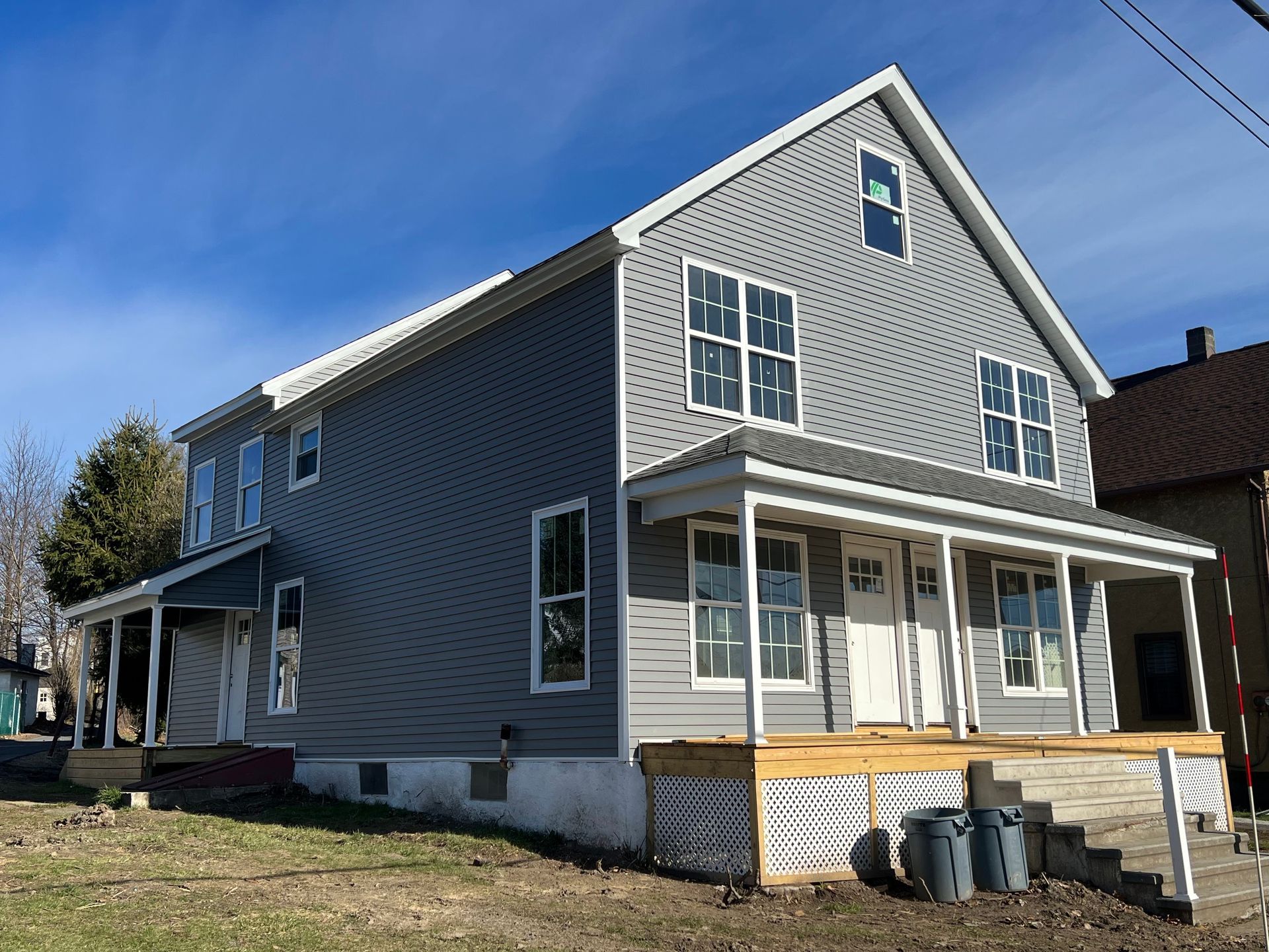 A two-story house with grey siding and a front porch, under construction in a residential setting on a sunny day.