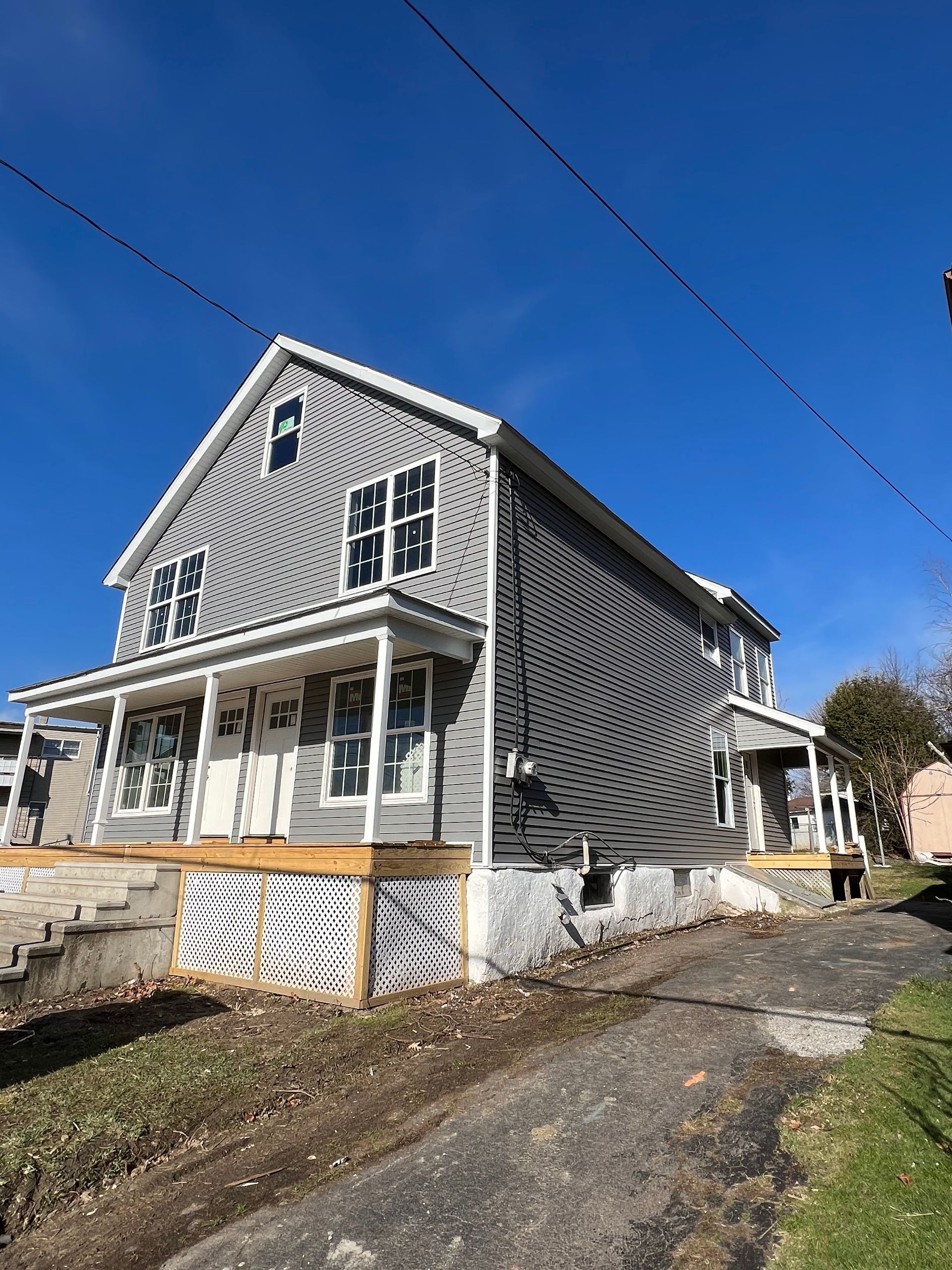 A two-story grey vinyl-sided house with a white front porch, lattice skirting, and a driveway under a clear blue sky.
