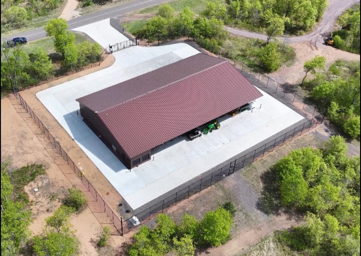Aerial view of a dark building with a reddish-brown roof on a large, light-colored concrete pad surrounded by a fence.