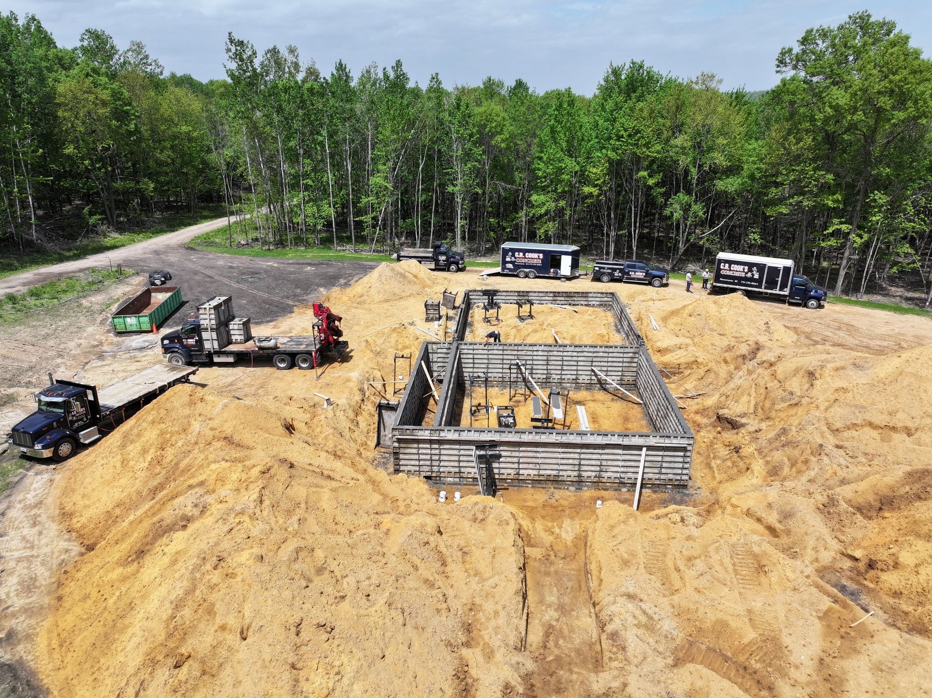 Construction site with foundation, dirt piles, and trucks in a wooded area.