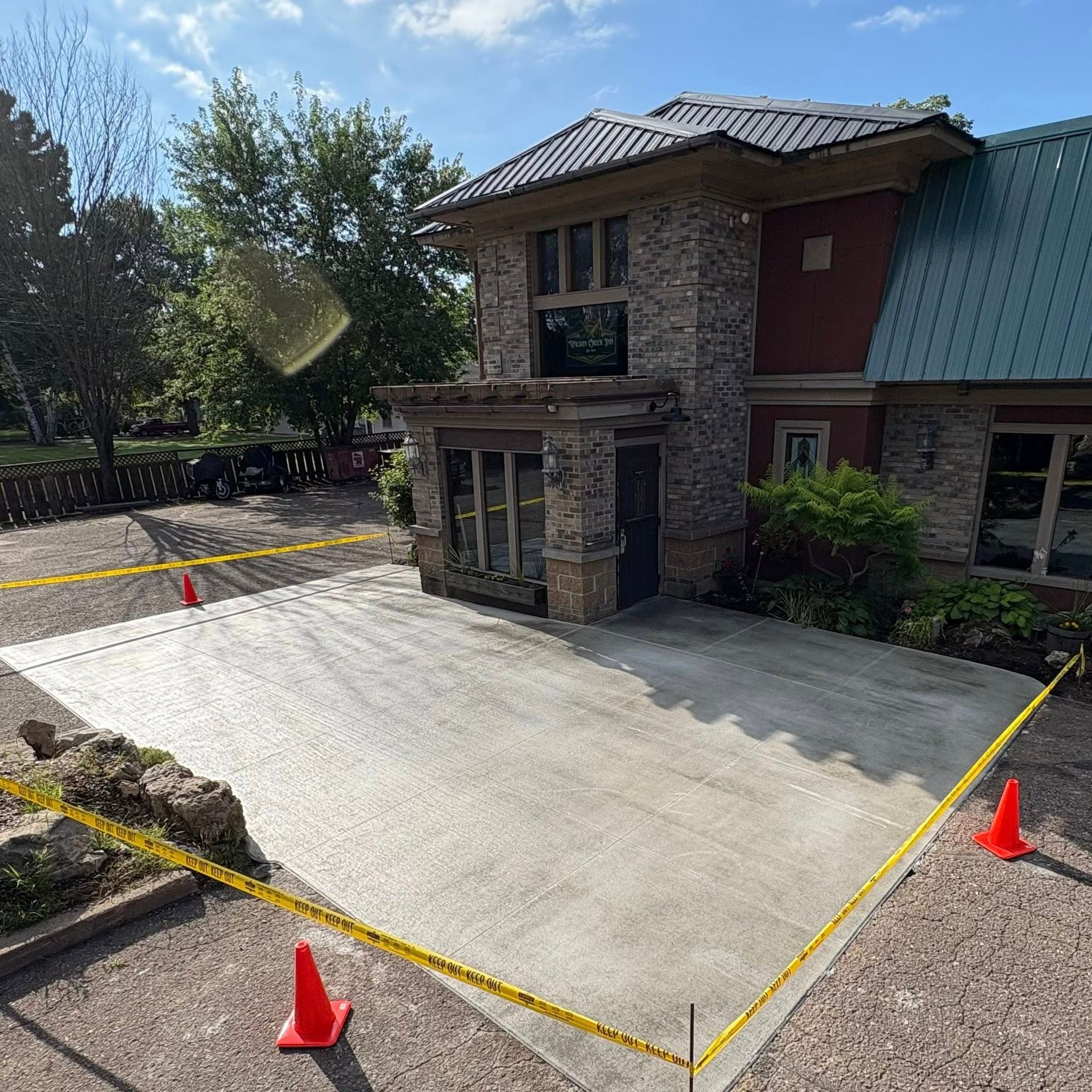Newly poured concrete driveway in front of a two-story building, marked with yellow caution tape and orange cones.