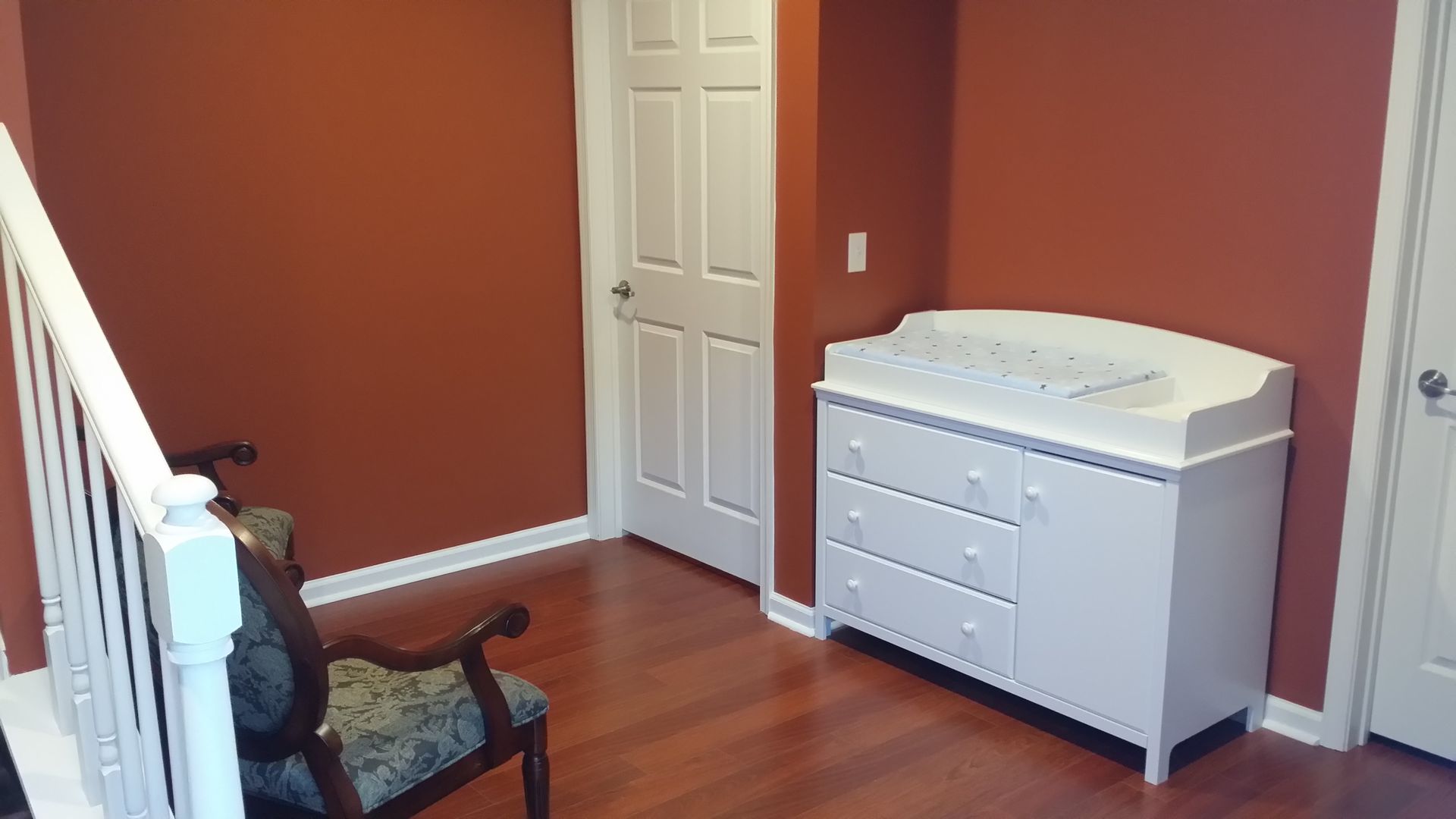Small bedroom with red walls, wooden floor, white dresser, and a closed door beside a staircase.