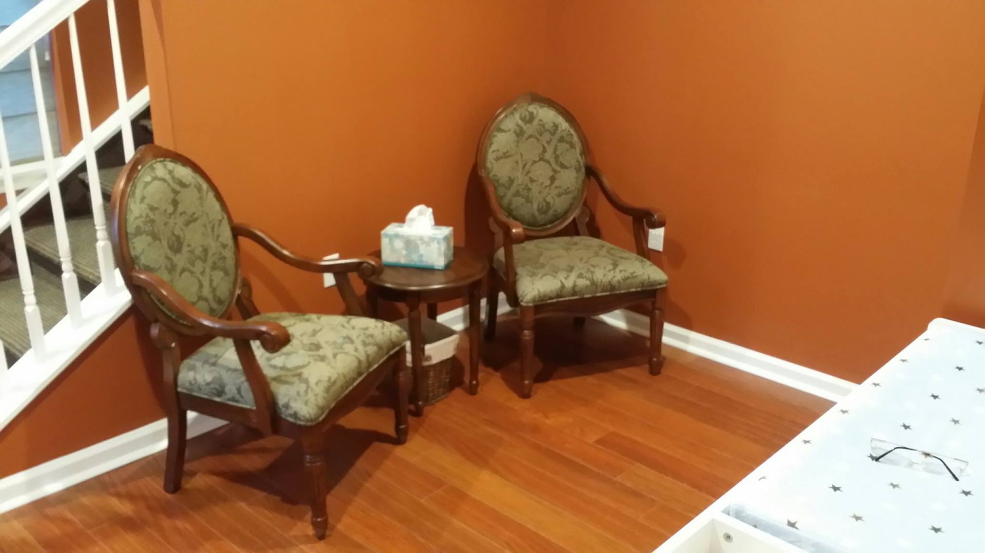 Two wooden chairs with green cushions beside a small side table in a room with orange walls and wood floor