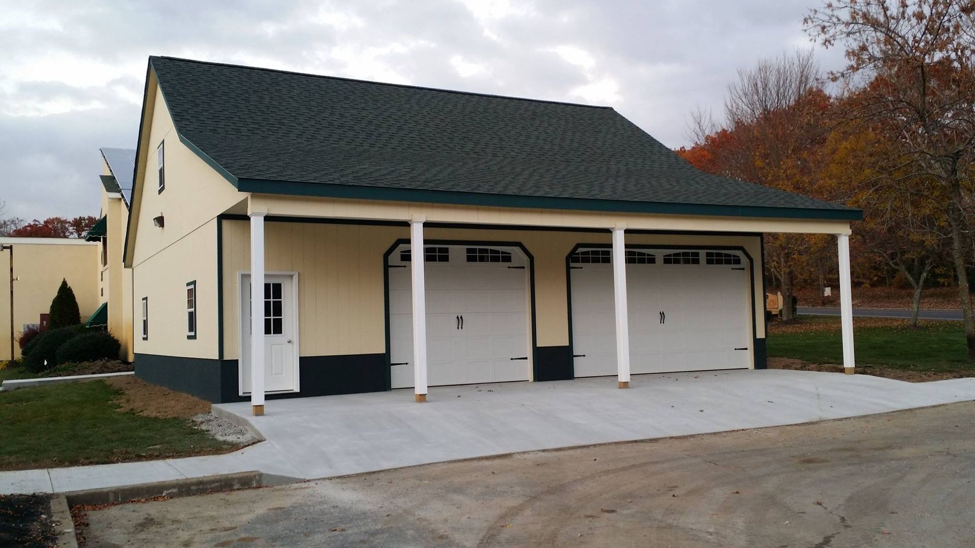 Detached beige double garage with green roof and white doors on a gravel driveway in autumn.