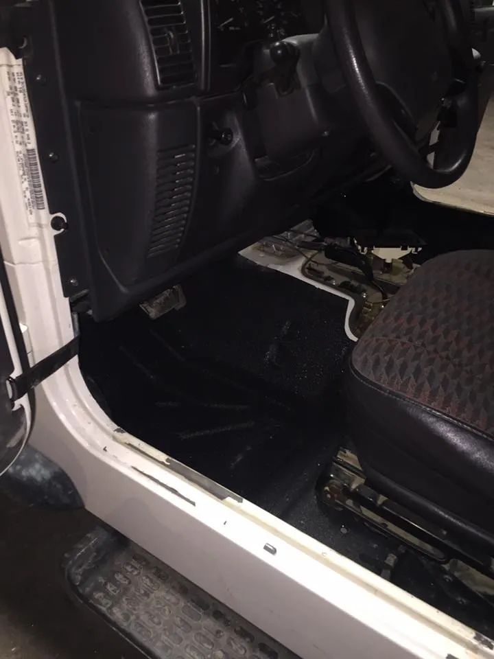 Interior of a white Jeep Wrangler. Black floor mats and seat. Black dashboard and steering wheel. Door is open.