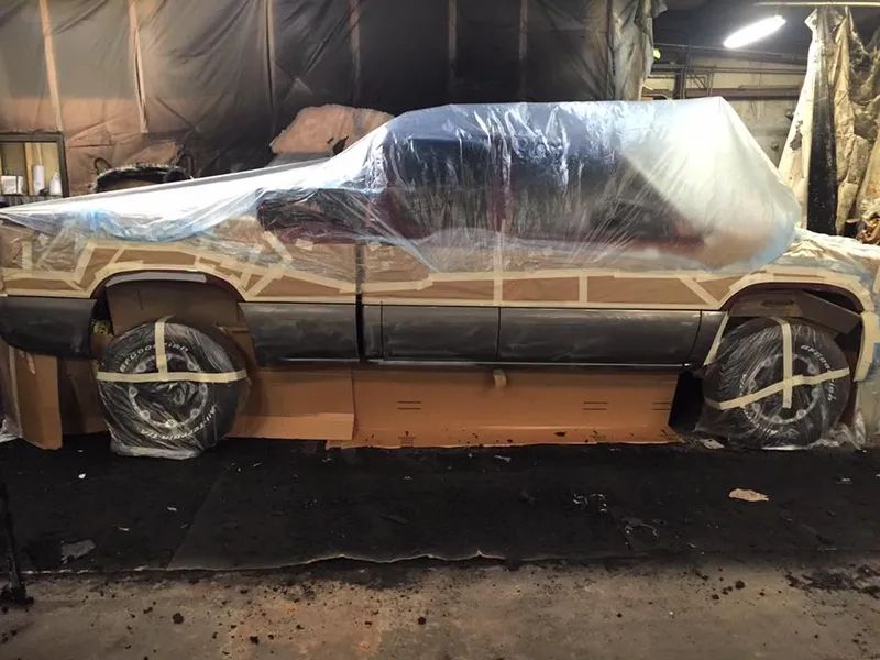 Truck undergoing bodywork, masked with tape and plastic sheeting. Wheels taped to cardboard base in a workshop.