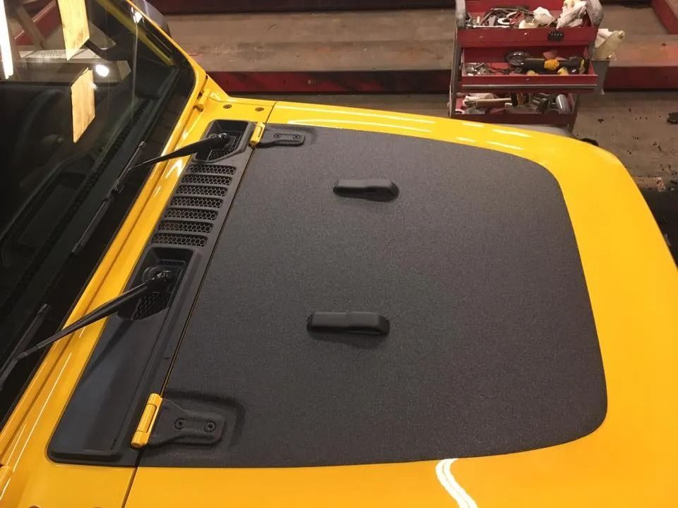 Yellow Jeep hood with black textured panel, two latches, windshield wipers, and a tool cabinet in the background.