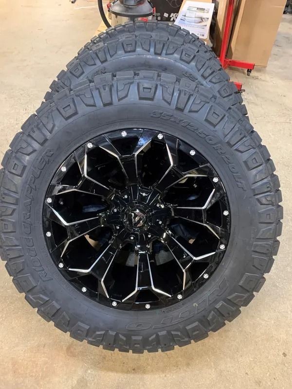 Black and white custom truck wheels with off-road tires, stacked on a garage floor.