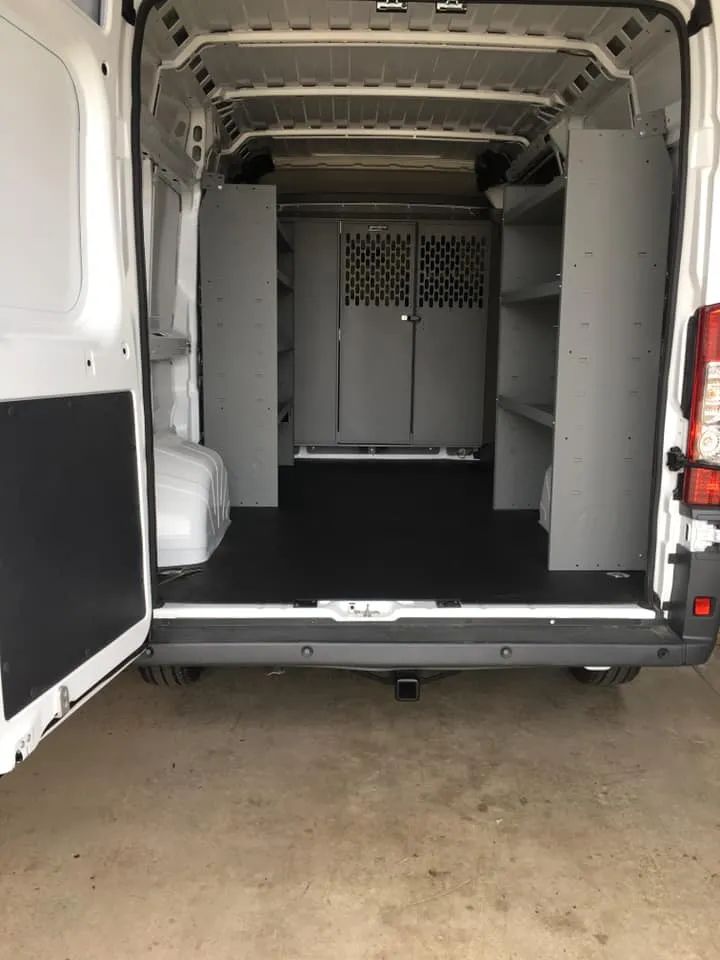 Interior view of a white cargo van with gray shelving and a dark floor. Rear doors are open.