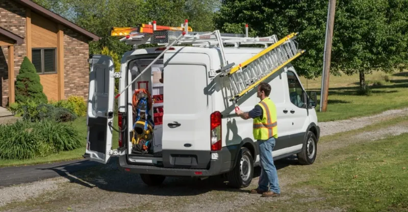 Man with a high-visibility vest removing a ladder from a work van, parked on a driveway with the back door open.