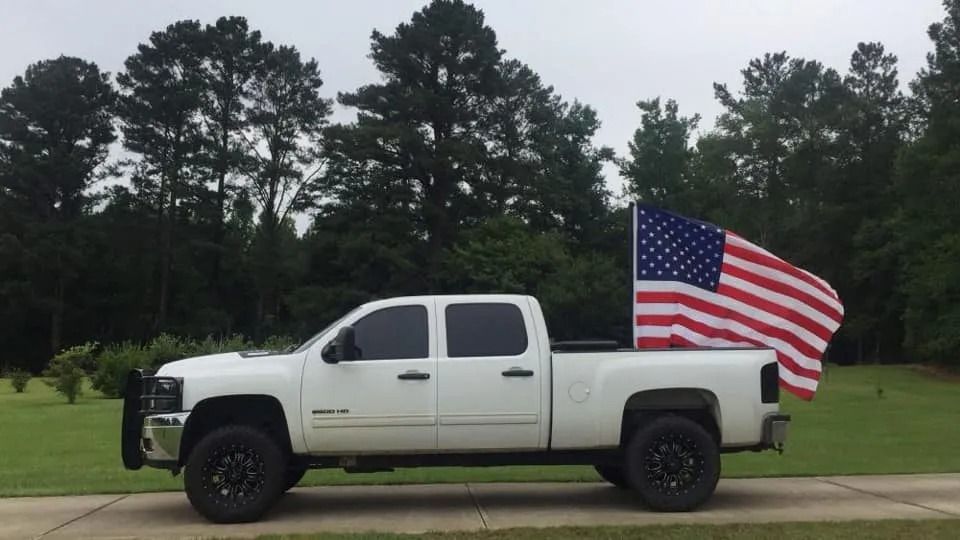 White pickup truck with American flag in the bed, parked on a paved road with trees in the background.