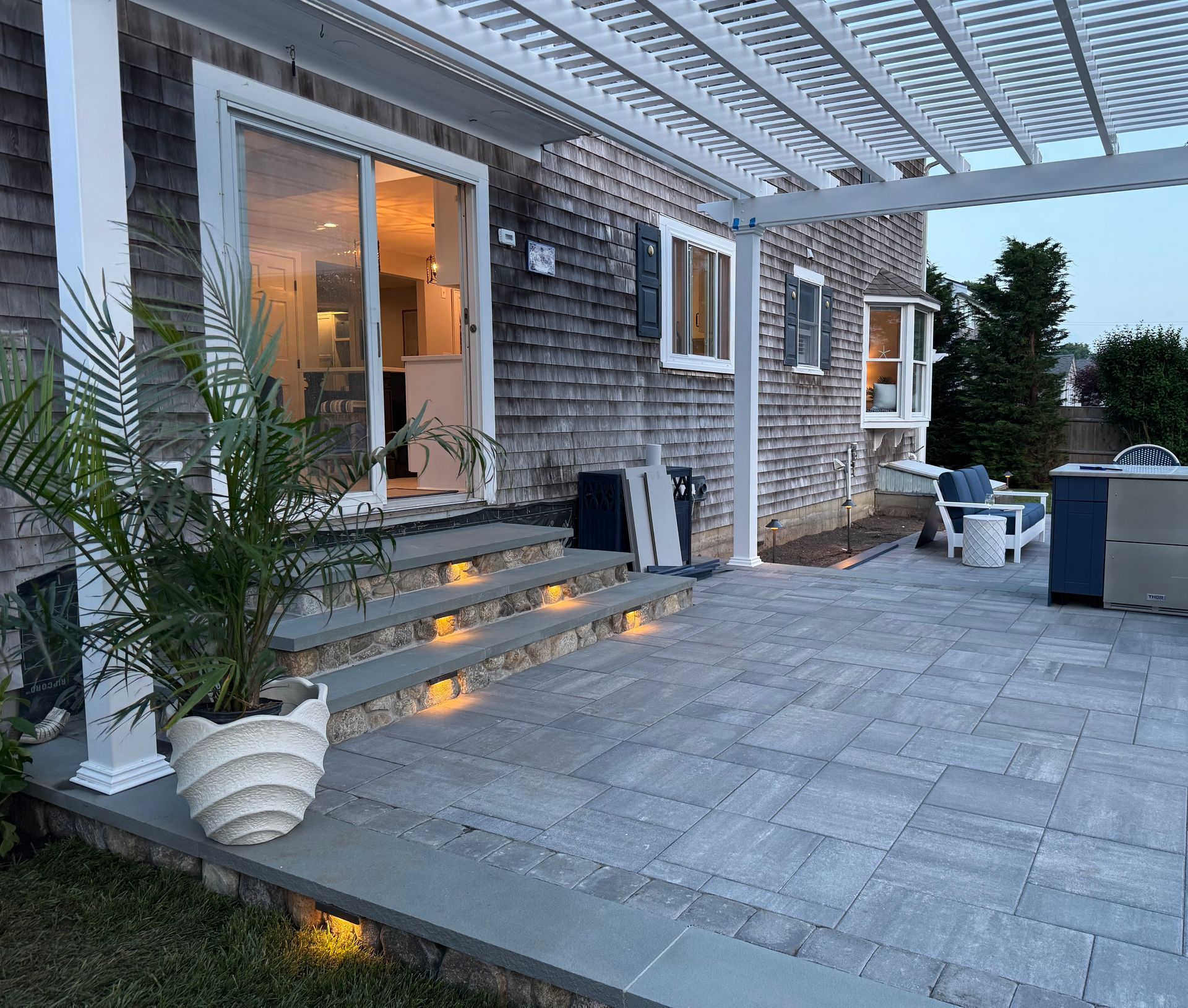 Modern backyard patio with stone steps, warm under-step lighting, pergola, and potted plants by a house.