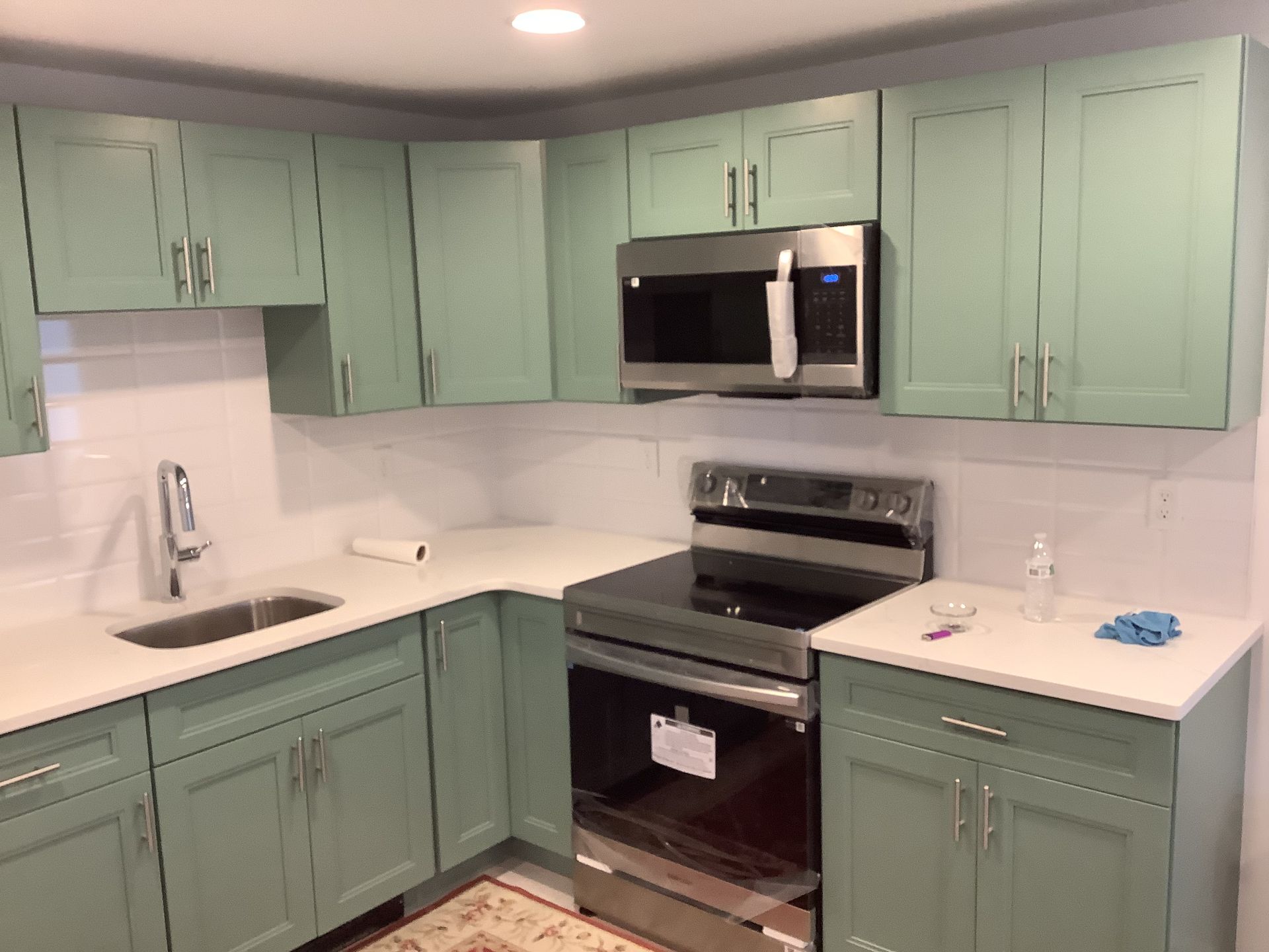 Green kitchen cabinets, white countertops, stainless steel appliances, white backsplash.