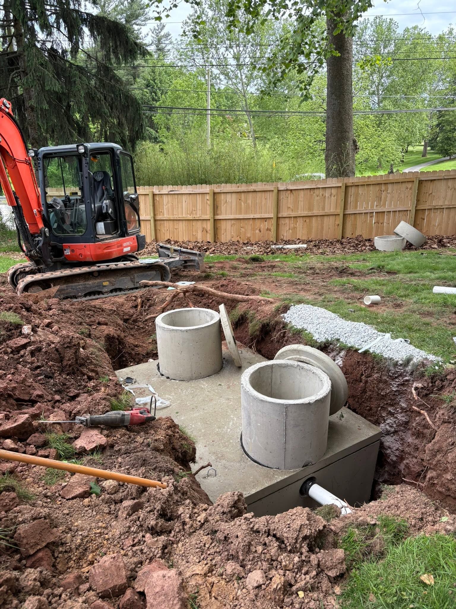 Backyard excavation with concrete utility pit and two gray pipe openings beside a mini excavator.