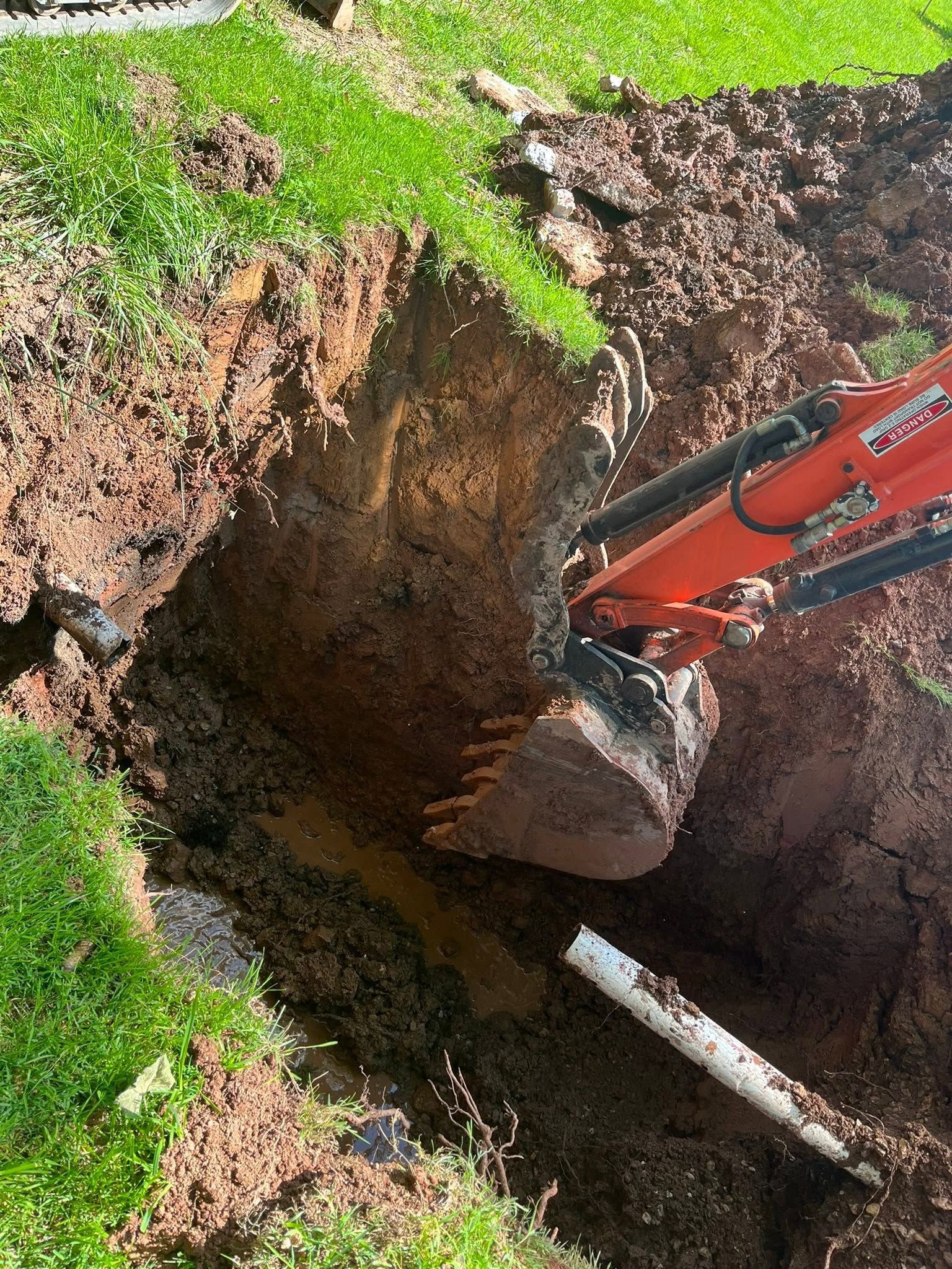 Excavator digging a deep hole in a grassy yard, exposing soil and roots