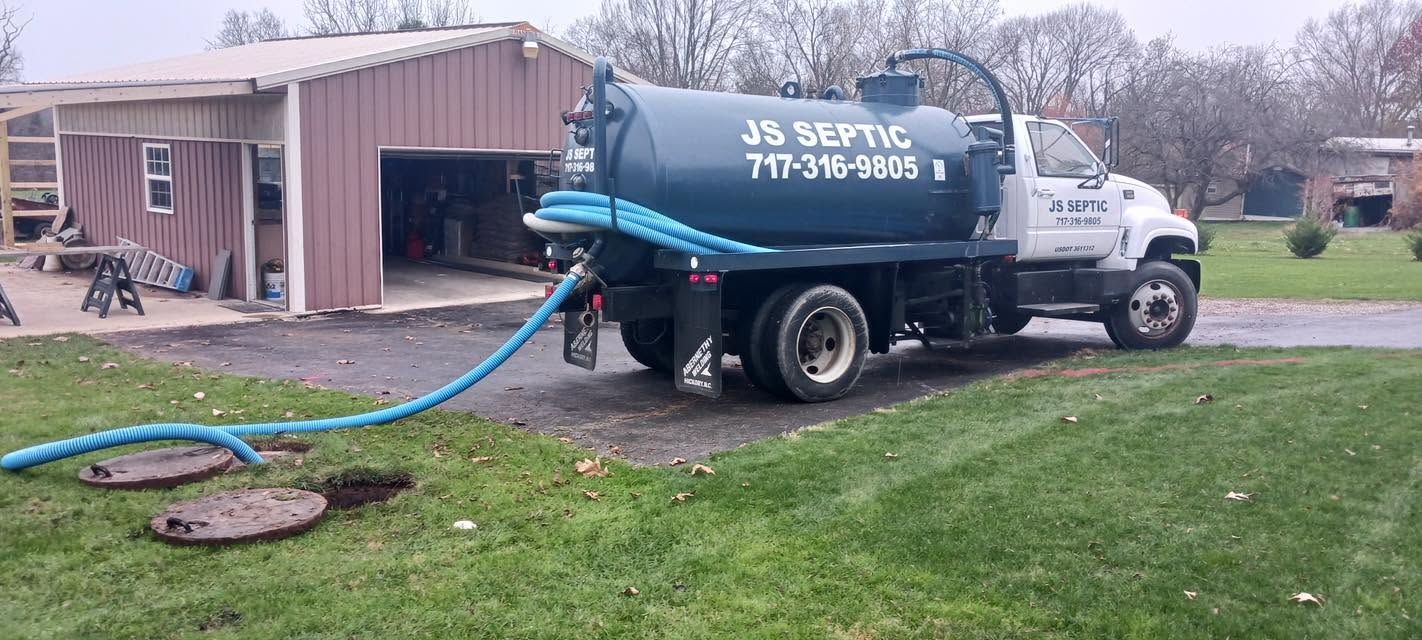 Blue septic tanker truck parked by a rural building, pumping out a tank through a hose.