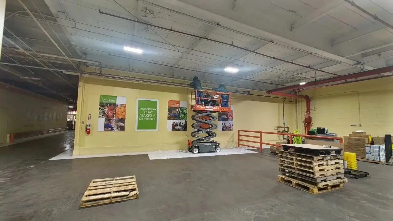 Warehouse interior with lift equipment, pallets, and posters on a light yellow wall. Concrete floor, overhead lights.