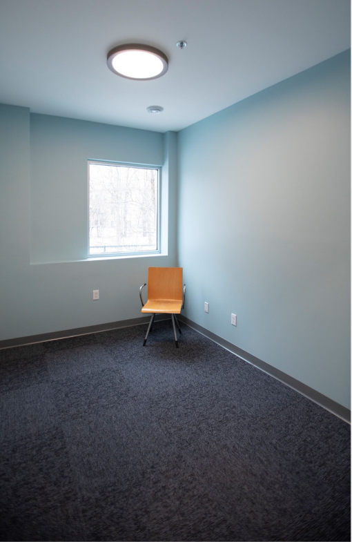 Empty light blue room with dark carpet, small window, and a single wooden chair near the corner.